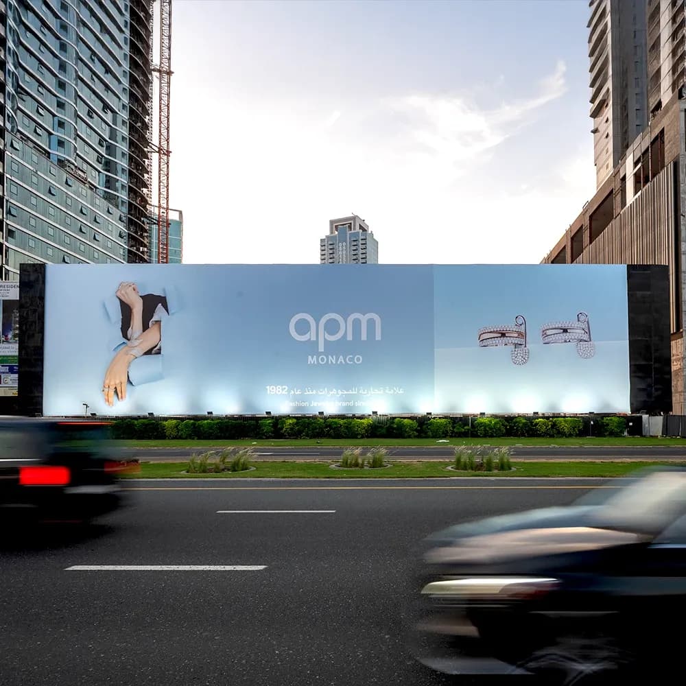 APM Monaco large-format roadside billboard advertising along a busy Dubai highway — premium static hoarding showcasing jewellery campaign with illuminated branding in an urban city setting.